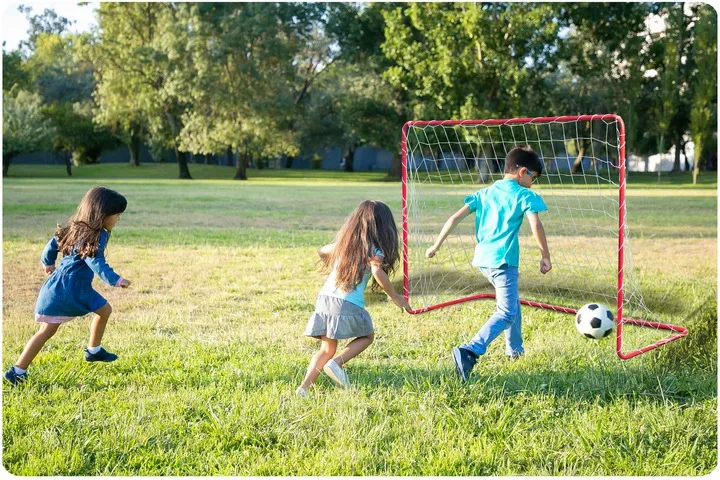 Bērni spēlē futbolu pie nelieliem vārtiem parkā ar bumbu pie vārtu malas