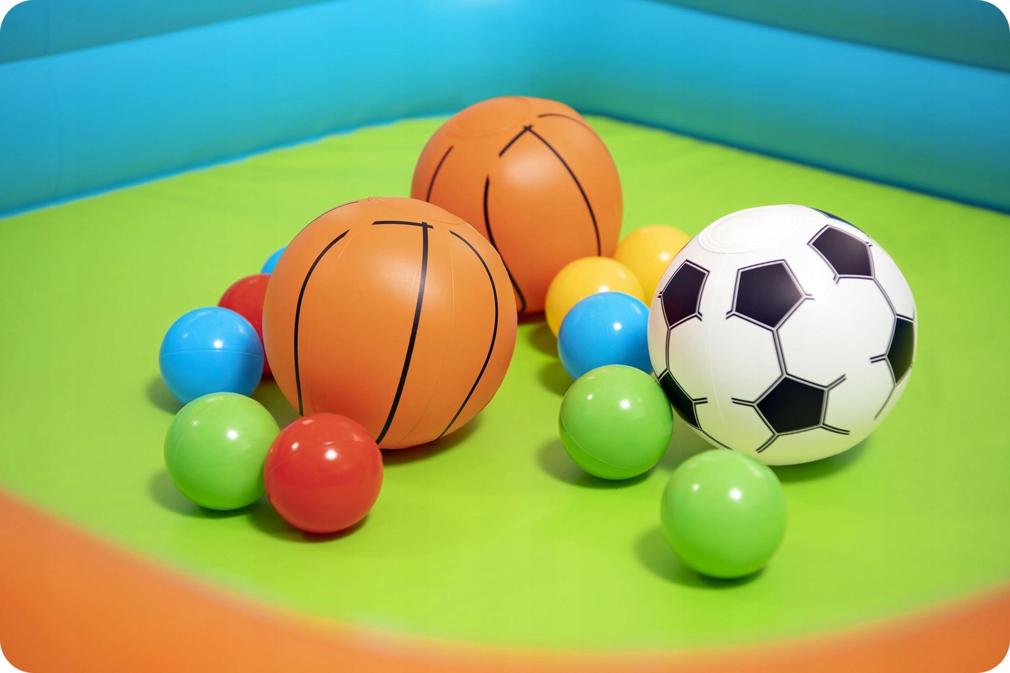 Tuvplānā redzamas rotaļu bumbas — divas basketbola bumbas, futbola bumba un krāsainas plastmasas bumbiņas uz zaļas pamatnes