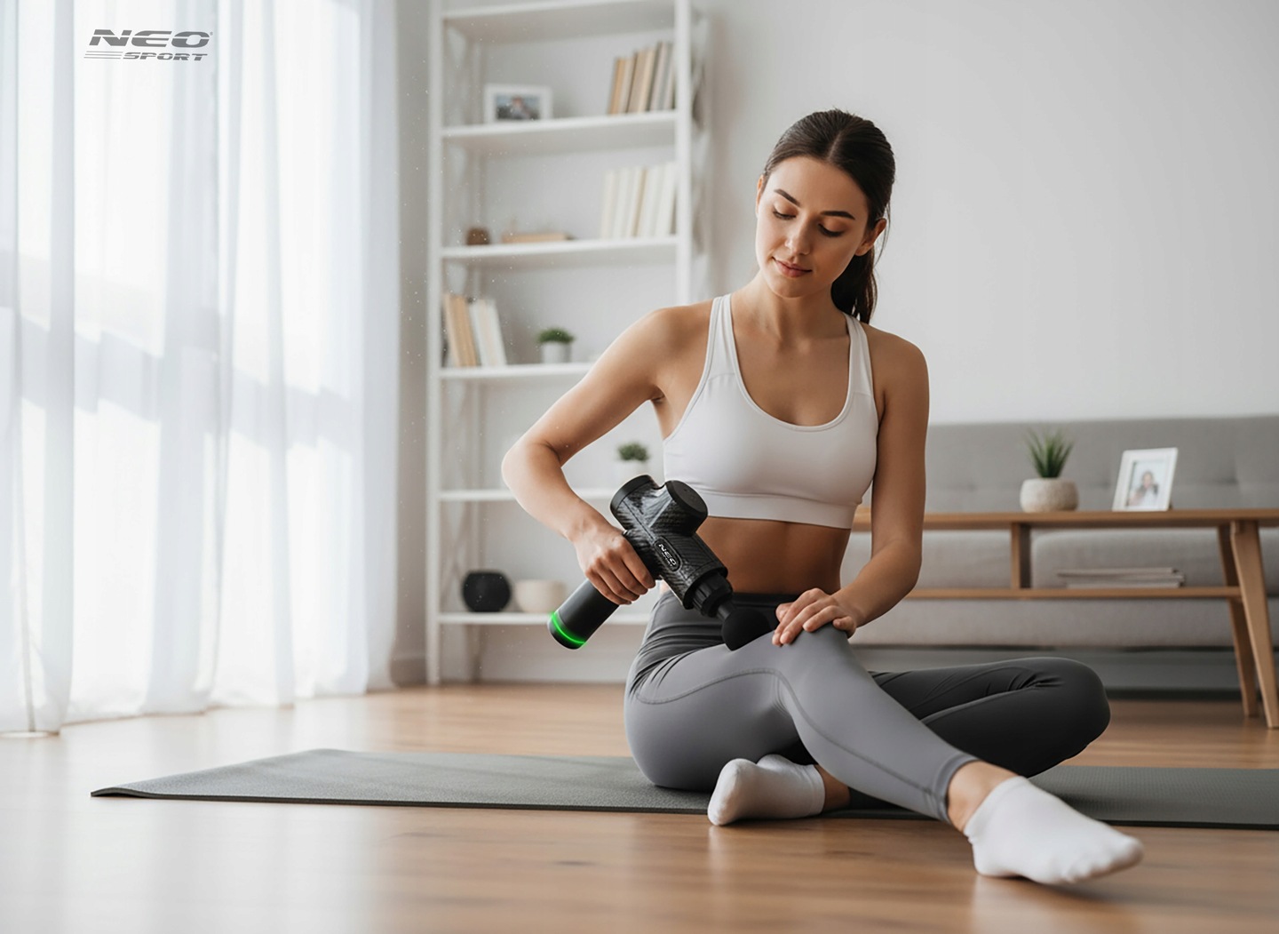 Sieviete uz jogas paklāja mājās lieto Neo-Sport masāžas pistoli, veicot relaksējošu masāžu
