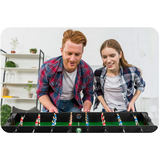 Divi cilvēki spēlē galda futbolu pie mājas galda ar redzamu laukuma malu un rokturiem