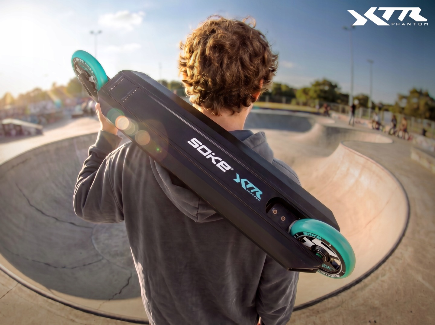 Braucējs nēsā SOKE XTR Phantom triku skrejriteni uz pleca pie skateparka, redzams deck apakšējais korpuss un tirkīza riteņi
