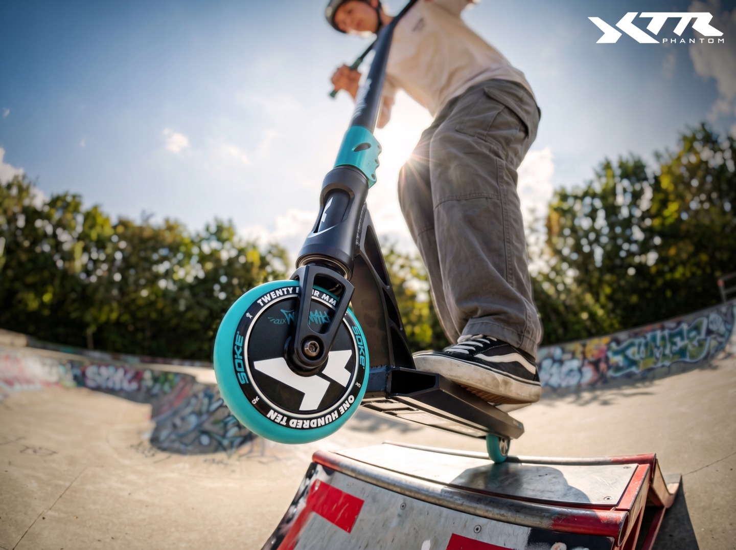 Braucējs ar SOKE XTR Phantom triku skrejriteni veic triku skateparkā, redzams zems kameras leņķis un priekšējais ritenis uz rampas malas