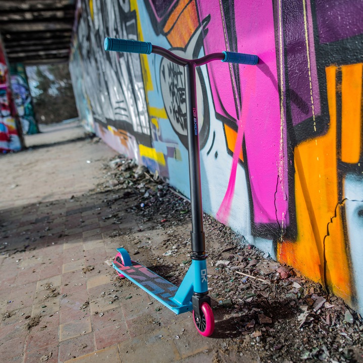 SOKE GO! triku skrejritenis pie grafiti sienas pilsētvidē ar turkīza un rozā krāsu akcentiem