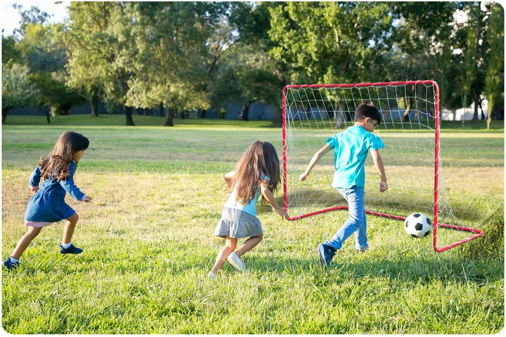 Futbola vārti parkā, bērni skrien uz bumbu pie sarkanā vārtu rāmja