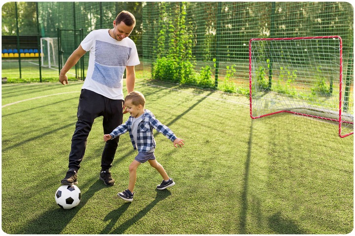 Futbola vārti sporta laukumā, pie vārtiem spēlējas pieaugušais ar bērnu un bumba