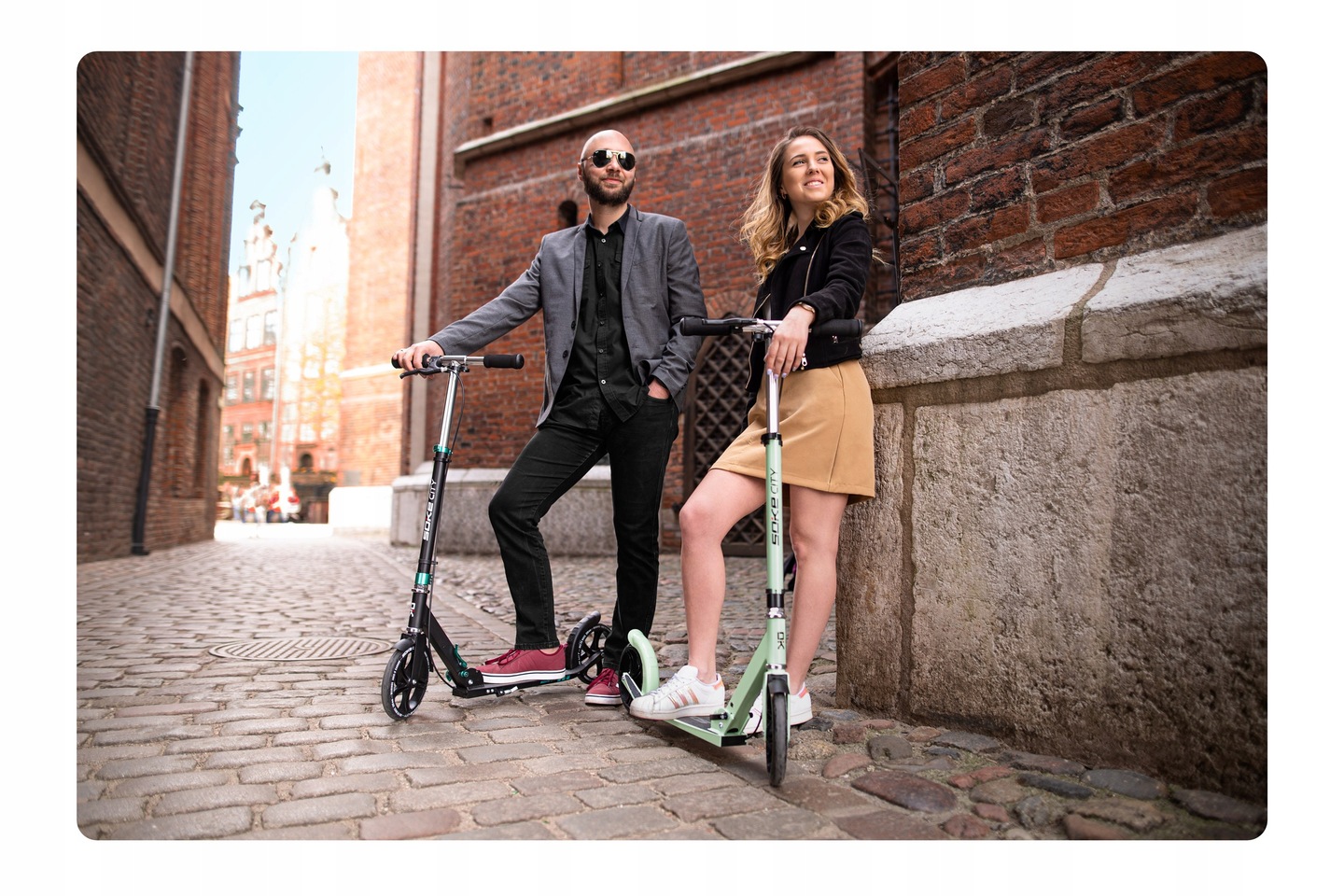 Divi cilvēki ar SOKE City skrejriteņiem stāv uz bruģētas pilsētas ielas pie ķieģeļu sienas