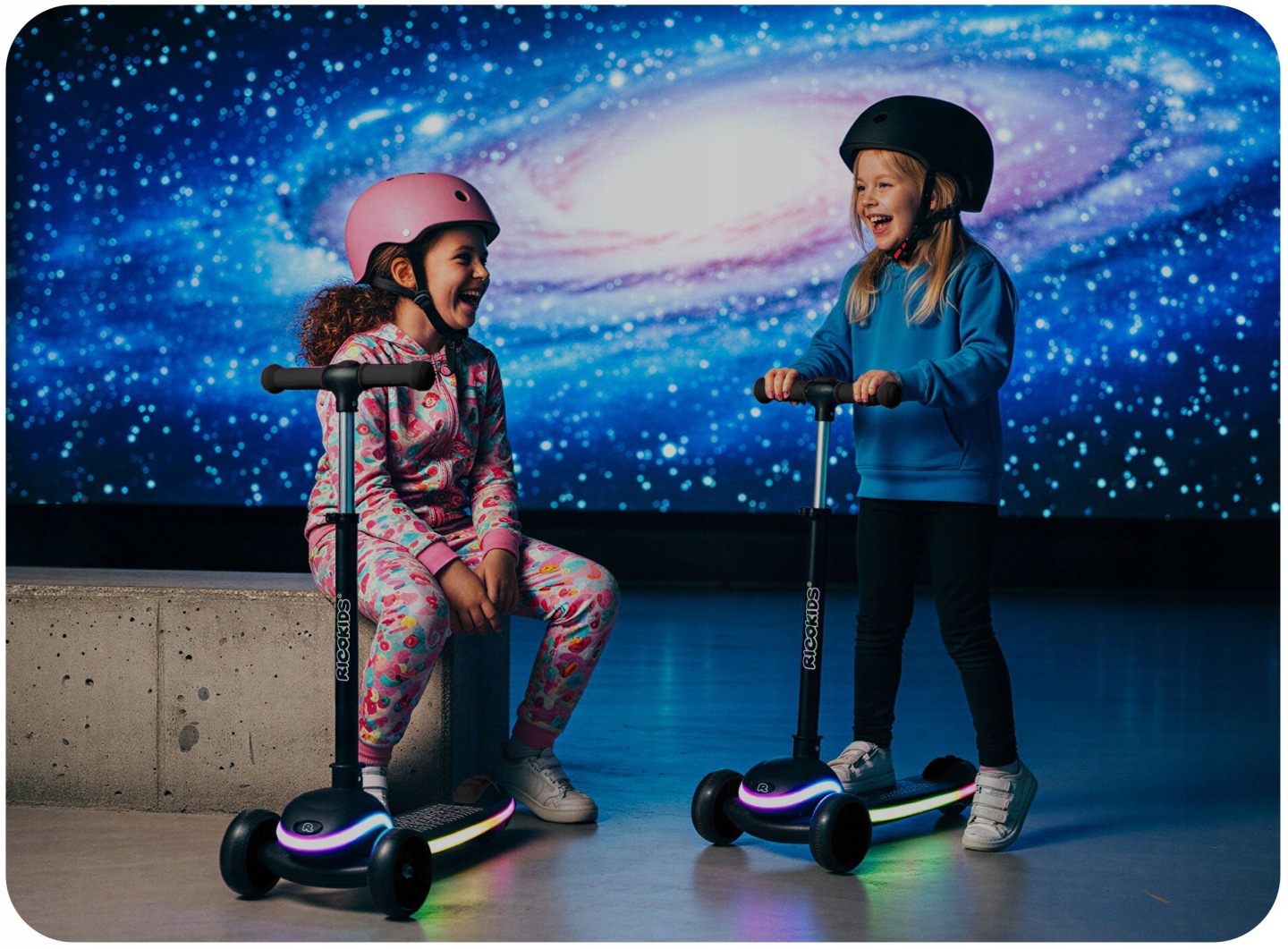 Divi bērni ar Ricokids Leddi trīsriteņu skrejriteņiem pie krāsaina, kosmosa tematikas fona