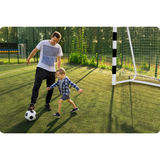 Tēvs un bērns spēlē futbolu pie vārtiem uz mākslīgā seguma laukuma